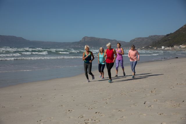 Group of women running on a beach on a sunny day, enjoying exercise and smiling. Perfect for promoting fitness, active lifestyle, wellness, and outdoor activities. Ideal for use in health and fitness campaigns, travel brochures, and advertisements focusing on group activities and friendship.