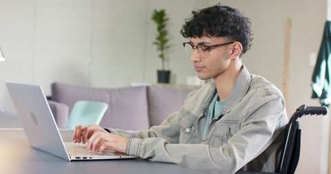 Indian young man in wheelchair typing on laptop in modern home workspace for remote work