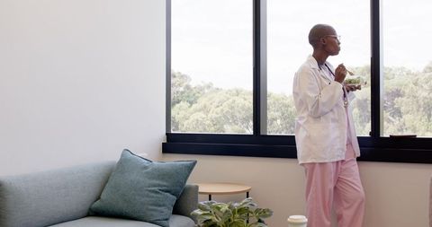Nurse taking break at window eating salad in calm modern clinic lounge with stethoscope