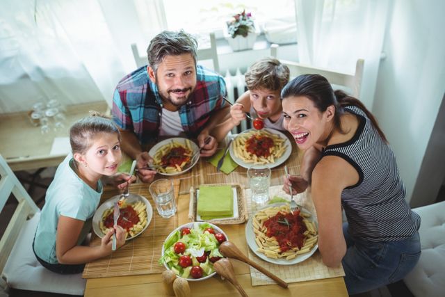 Family sitting around table, enjoying time eating pasta meal together at home. Suitable for content related to family bonding, home life, dining experiences, and casual meals. Ideal for use in family-oriented advertisements, cooking blogs, or lifestyle articles.
