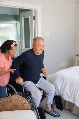 Senior asian caregiver assisting man transferring from wheelchair to bed at home
