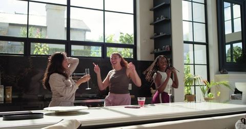 Sunlit diverse women laughing and dancing by white kitchen island at casual gathering