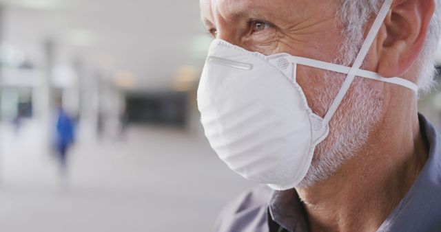Senior man wearing a white protective face mask in a public indoor space with a blurred background. The image is useful for illustrating health safety measures, medical protocols, elderly care, and public health awareness. Can be used in health-related articles, medical information pamphlets or websites highlighting the importance of wearing masks.