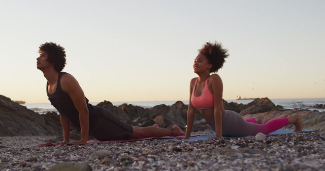 Young couple practicing yoga on a beach during sunset. They are stretching on their mats, showing physical strength and focus. Ideal for uses in wellness, yoga classes, outdoor exercise promotions, and lifestyle blogs.