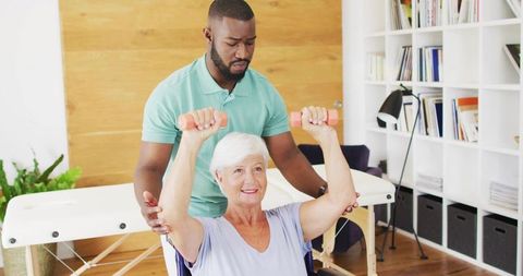 Senior woman raising dumbbells while caregiver assisting during rehabilitation session
