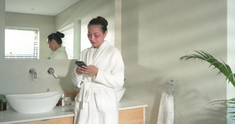 Asian woman using smartphone in modern bathroom wearing white robe and pink eye patches