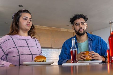 Youthful diverse couple sitting at kitchen counter eating burgers and drinking soda