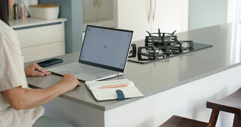 Mature woman working remotely at kitchen island with laptop and notebook, bright minimalist home off