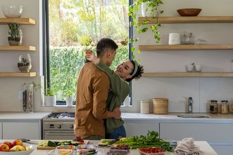Couple embracing while preparing fresh vegetables in sunlit modern kitchen with plants and smiles
