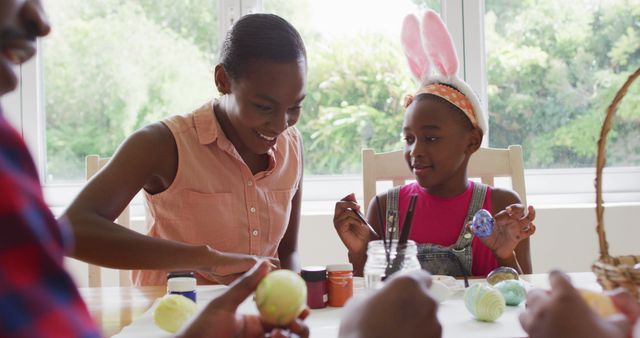 A mother and daughter are painting Easter eggs together at home. The daughter is wearing bunny ears while holding a painted egg. This scene can be used for content related to holiday activities, family bonding, creative projects, or Easter celebrations.
