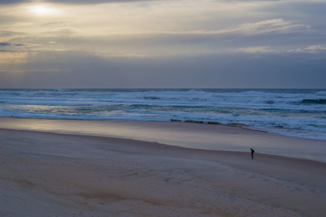 A solitary person walks along a vast, empty beach in the early morning as the sun rises over the horizon, casting a tranquil light on the sand and waves. Ideal for themes of solitude, reflection, nature, peaceful moments, and relaxation.