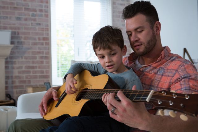 Father and young son bonding through music as they play acoustic guitar together in a cozy living room. Ideal for themes related to family activities, music education, father-son relationships, home life, and parenting.