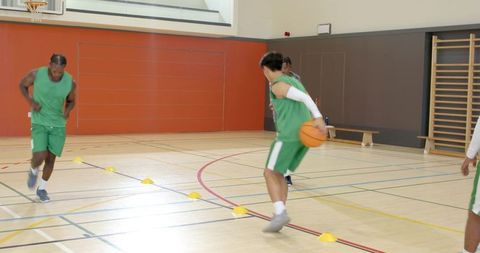 Diverse male basketball players practicing cone dribbling and passing drill in indoor gym