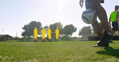Striking soccer ball during training drill with yellow mannequins on sunny artificial turf