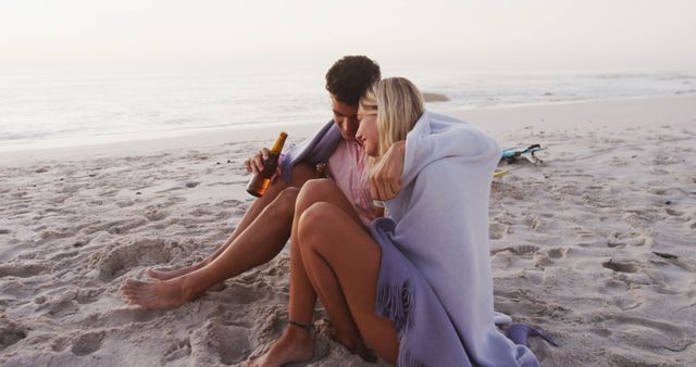 Happy diverse couple covered with blanket embracing and drinking beer on beach. Lifestyle, relaxation, nature, free time and vacation.