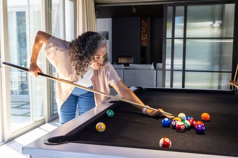 Woman playing pool in modern sunlit living room leaning over black felt table