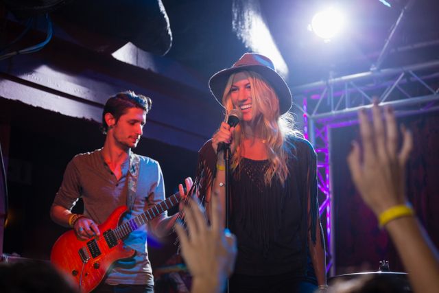 Singer performing on stage in a nightclub with guitarist in the background. Audience members raising hands, enjoying live music. Ideal for use in promotions for live music events, nightlife advertisements, and entertainment industry content.