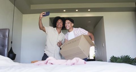 Young friends moving home smiling while taking selfie with donation box in bright bedroom