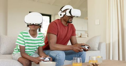 Father and Son Wearing VR Headsets Playing Video Games on Sofa at Home