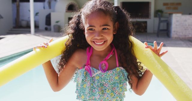 Happy biracial girl is holding a yellow pool noodle, smiling brightly while standing at the edge of a backyard swimming pool. The scene suggests leisure and joy as a child enjoys summer activities at home, making this suitable for use in depicting recreational fun, family moments, and outdoor activities.