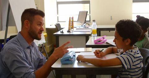 This depicts a teacher actively engaging with a young schoolboy in a bright, modern classroom setting. They are focused on a task requiring collaboration and attention, conveying themes of education and personal mentoring. Suitable for educational websites, learning blogs, or promotional materials for schools, highlighting focused, one-on-one instruction.