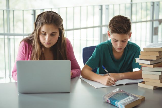 Two school kids are studying in a library. One is using a laptop while the other is writing in a notebook. This image can be used for educational content, school promotions, academic articles, and learning resources.