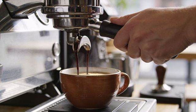 Barista extracting espresso into steaming brown cup, crema pouring from portafilter in cafe