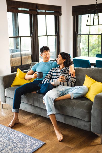 Multiracial gay couple sitting on a couch in a cozy living room, holding coffee cups and engaging in conversation. Ideal for use in articles or advertisements focusing on LGBTQ relationships, home lifestyle, or casual bonding moments. Perfect for promoting inclusivity, love, and modern living.