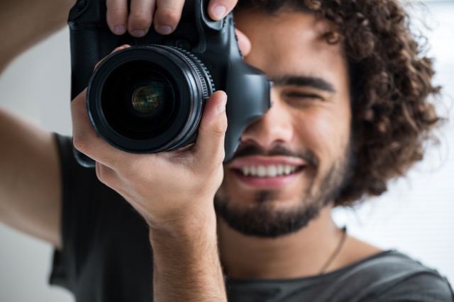 Image shows a happy male photographer with curly hair taking a picture with a digital camera in a studio. Ideal for usage in content related to photography, hobbies, professional services, creative industries, and lifestyle blogs showcasing behind-the-scenes moments.
