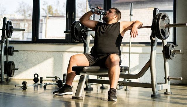 Man drinking water on bench press between sets in sunlit gym, resting after heavy lift
