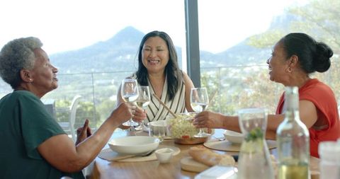 Diverse senior women sharing salad and wine on sunlit terrace while laughing and toasting