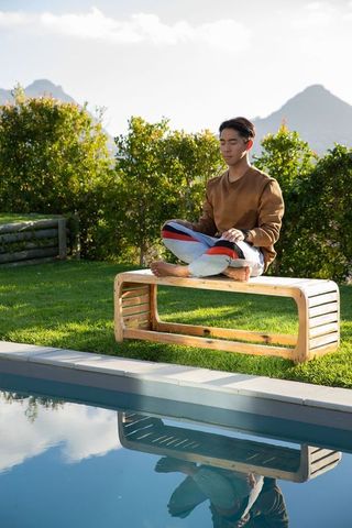 Asian man meditating on wooden bench beside pool with mountain reflection and garden
