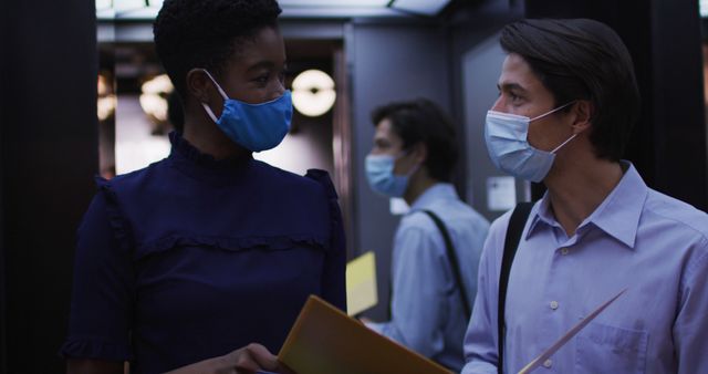 Picture of colleagues wearing protective masks while having a conversation in an office elevator. This image can be used to depict workplace safety, COVID-19 measures, business continuity, and office communication during the pandemic.