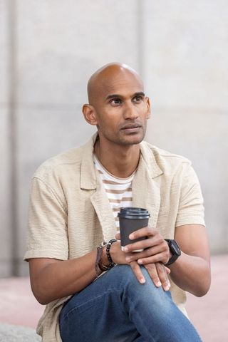 Contemplative mid-40s man holding coffee cup sitting urban plaza wearing beige overshirt