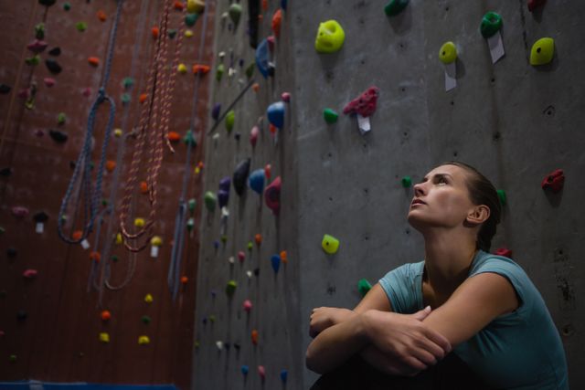 Female climber sitting and looking up thoughtfully in an indoor climbing gym. Colorful climbing holds and ropes are visible on the wall. Ideal for use in articles or advertisements related to fitness, rock climbing, mental focus, and sports training.