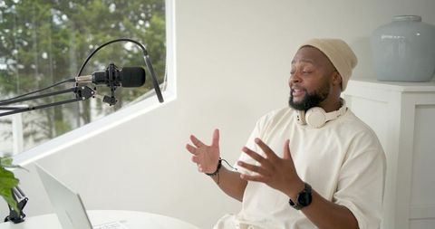 Black podcaster recording in minimalist home studio, gesturing toward laptop during livestream