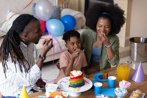 African american family celebrating birthday, eating rainbow cake at cozy living room party