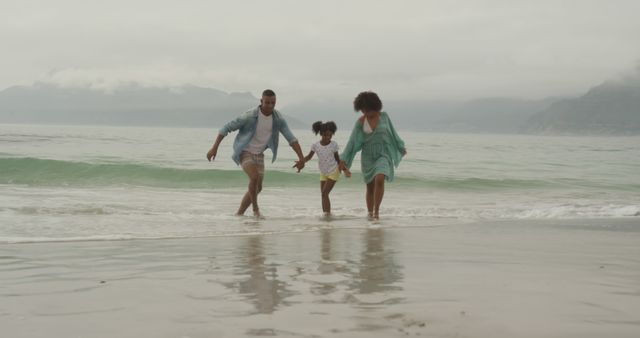 This joyful scene depicts a happy family enjoying their time together at the beach, running through shallow ocean water on a cloudy day. The child is holding hands with the parents, conveying a strong sense of bonding and playful activity. This photo is perfect for use in advertisements, travel promotions, family-focused marketing, and parenting blogs. It portrays themes of family time, parenting, and outdoor fun.