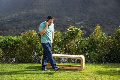 Man talking on phone walking past wooden bench on sunlit lawn with mountain view
