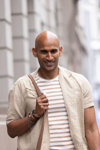 Smiling man walking urban street wearing striped tee and linen jacket carrying crossbody satchel
