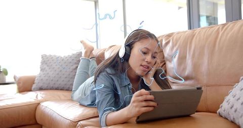 Teen lying on leather couch using tablet wearing headphones enjoying music in bright room