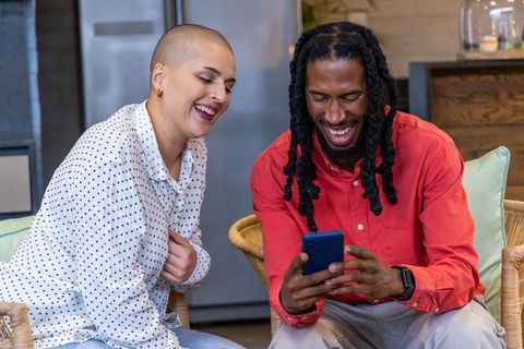 Multiracial couple laughing while looking at smartphone in cozy modern kitchen lounge