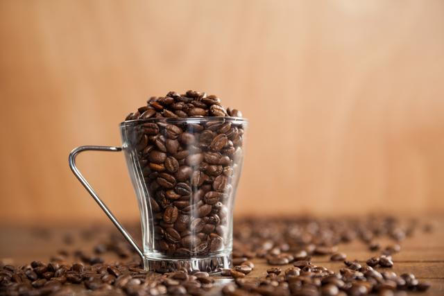 Glass cup filled with coffee beans on wooden table, perfect for illustrating coffee-related content, cafe promotions, or articles about coffee culture. Ideal for use in blogs, social media posts, and advertisements for coffee shops or coffee products.