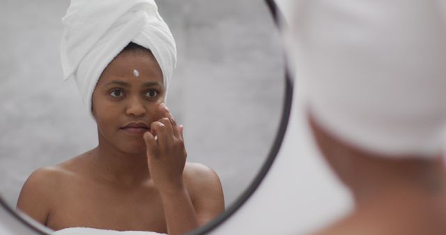 Woman with a white towel wrap around her head applying skincare cream on her face while standing in front of a mirror. Ideal for articles or promotions related to skincare routines, beauty products, self-care tutorials, and personal grooming techniques.