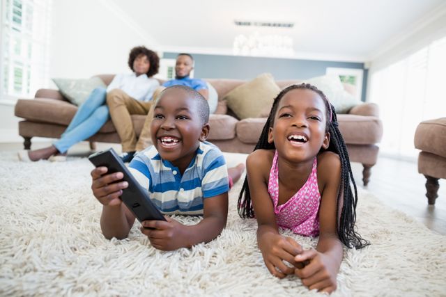 Siblings lying on a rug in a bright living room, watching television and smiling. Parents are sitting on the couch in the background, creating a warm family atmosphere. Ideal for use in advertisements, family-oriented content, and articles about home life, family bonding, and childhood leisure activities.