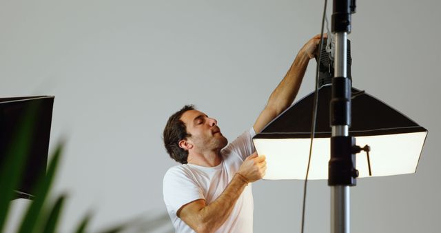 A middle-aged Caucasian man adjusts professional lighting equipment in a studio setting, with copy space. His focus and expertise suggest he could be a photographer or a lighting technician preparing for a photoshoot or film production.