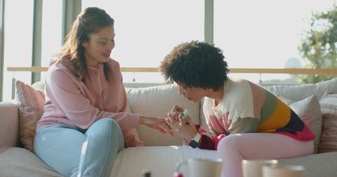 Friends giving manicure on sunlit couch — cozy at-home self-care and bonding moment