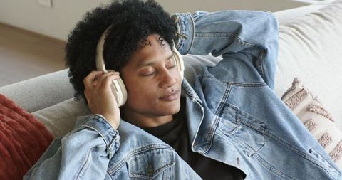 African american man relaxing on couch listening to music with headphones in denim jacket