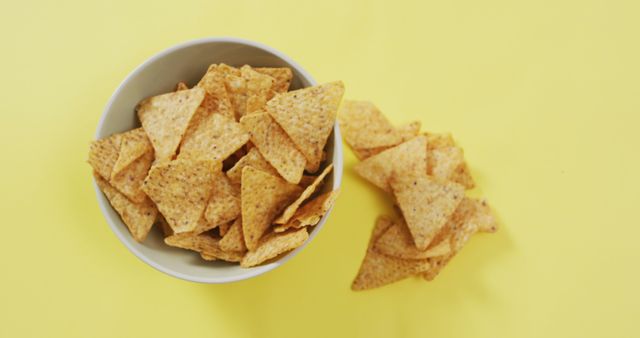 Bowl filled with crispy nachos and some chips scattered around on a bright yellow background. Ideal for food and beverage advertisements, party invitations, or casual dining promotions.
