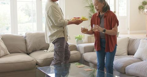 Asian senior couple sharing snacks and drinks in cozy sunlit living room with beige decor